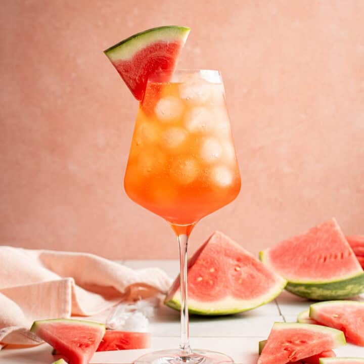 A wine glass filled with ice and Watermelon Aperol Spritz with a watermelon wedge.
