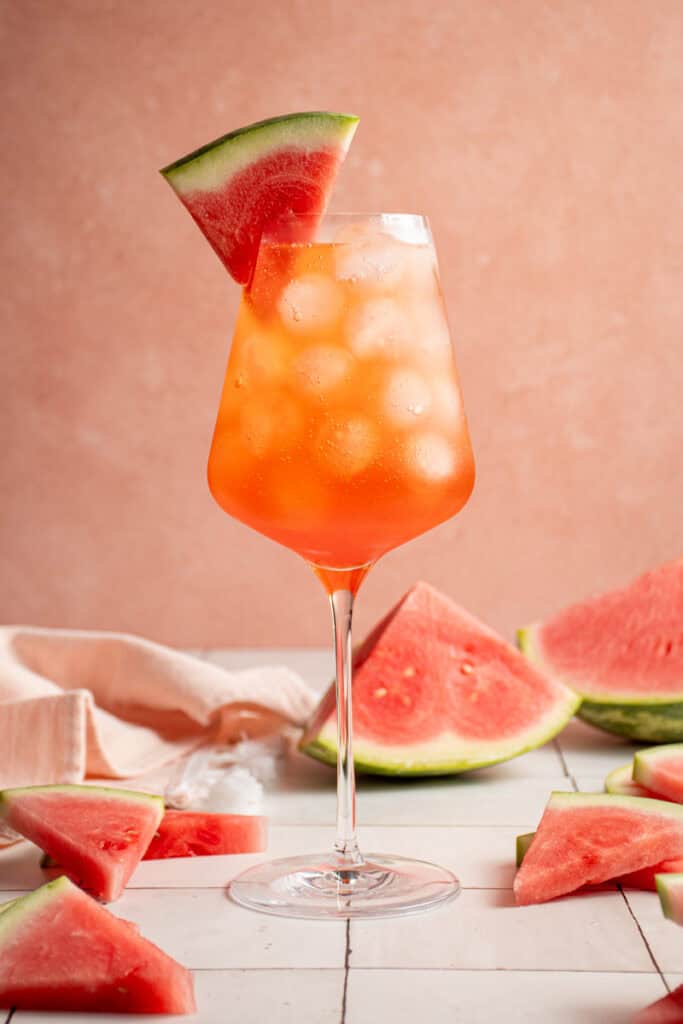 A wine glass filled with ice and Watermelon Aperol Spritz with a watermelon wedge.