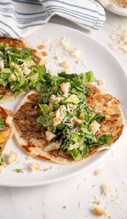 A plate with a smash chicken taco topped with chopped caesar salad with parmesan and crushed croutons.