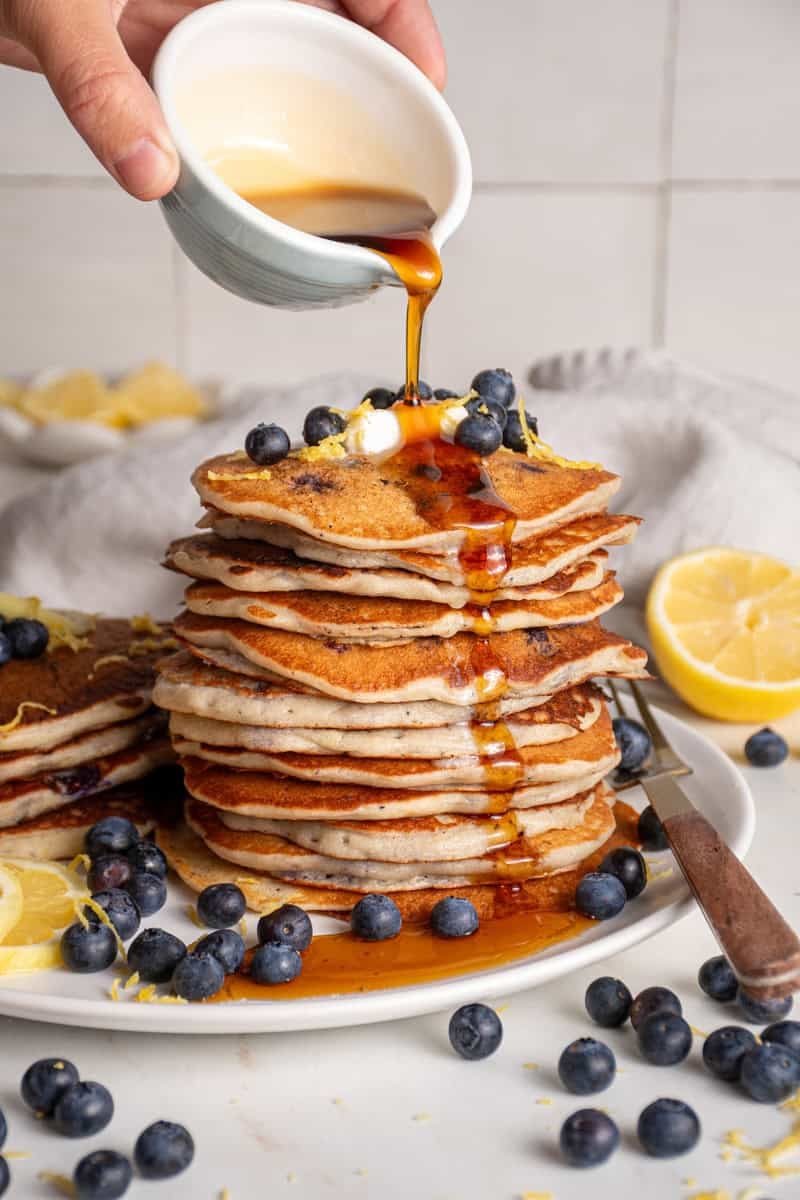 A stack of lemon blueberry pancakes with syrup pouring over the top.