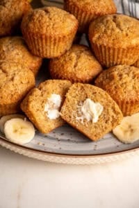 A plate filled with whole wheat banana muffins with one in the center cut in half.
