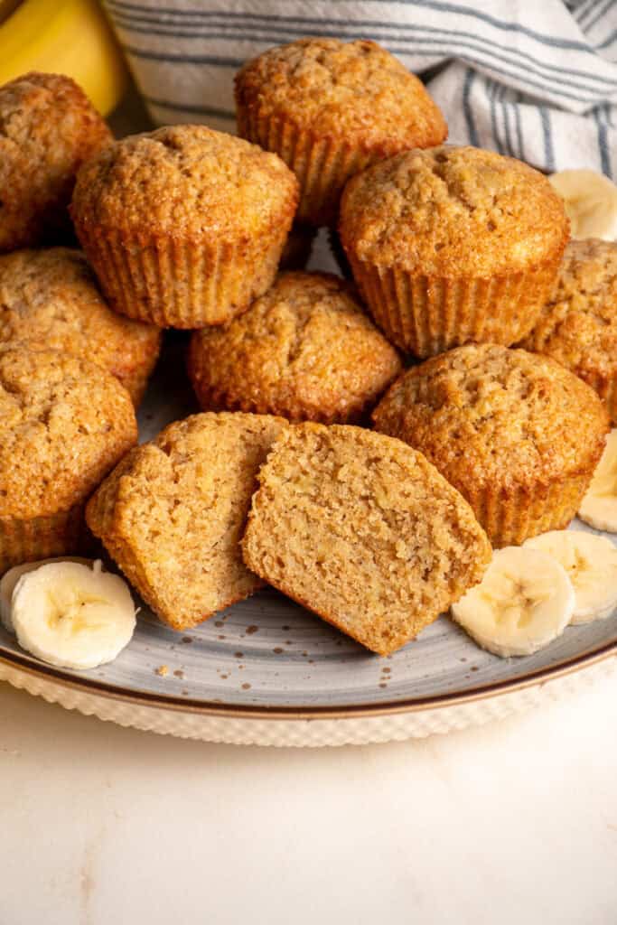 A plate filled with whole wheat banana muffins with one in the center cut in half.