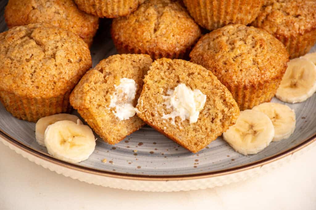 A plate filled with whole wheat banana muffins with one in the center cut in half.