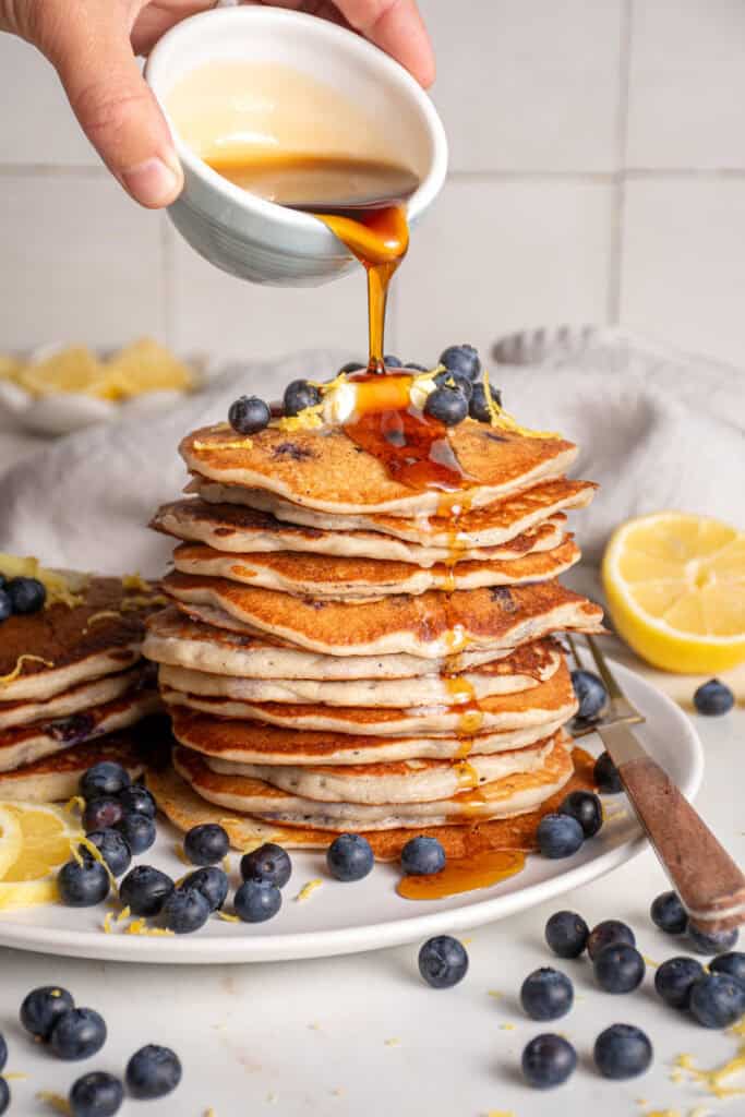 Pouring syrup over top a stack of freshly made lemon blueberry pancakes.