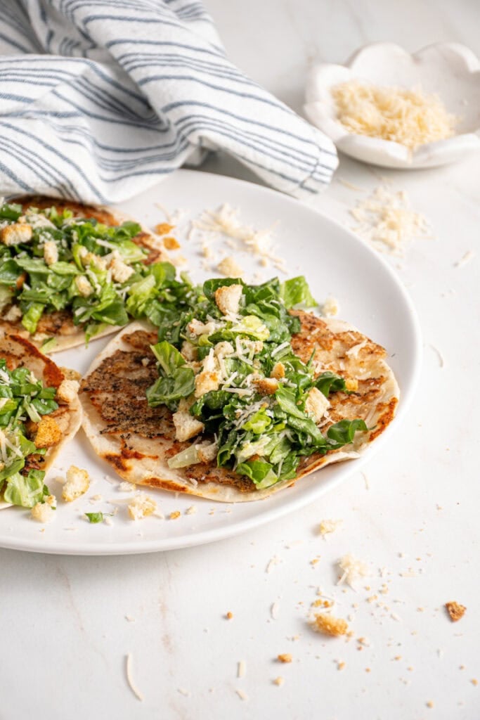 A plate with a smash chicken taco topped with chopped caesar salad with parmesan and crushed croutons.