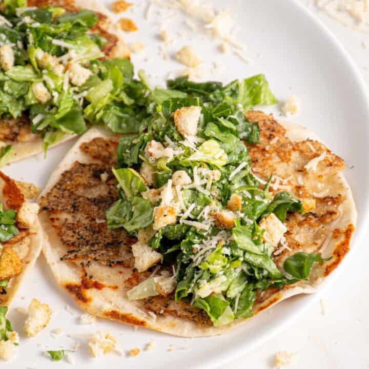 A plate with a smash chicken taco topped with chopped caesar salad with parmesan and crushed croutons.