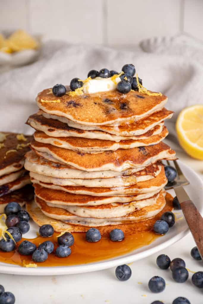 A stack of lemon blueberry pancakes with butter, syrup, and fresh blueberries.