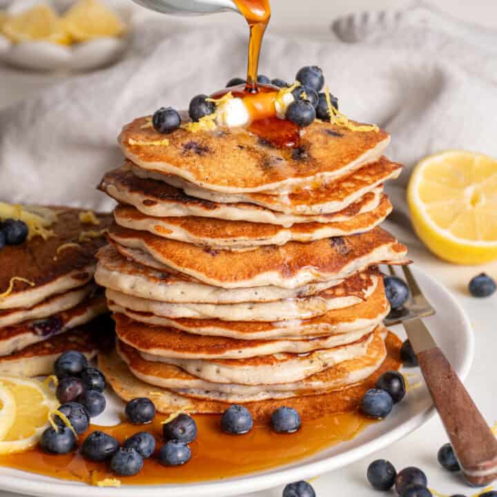 Maple syrup being poured onto a stack of lemon blueberry pancakes.