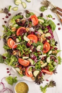 A large platter full of spring mix salad with tomatoes, cucumbers, red onion, walnuts, dried cranberries, and olives.
