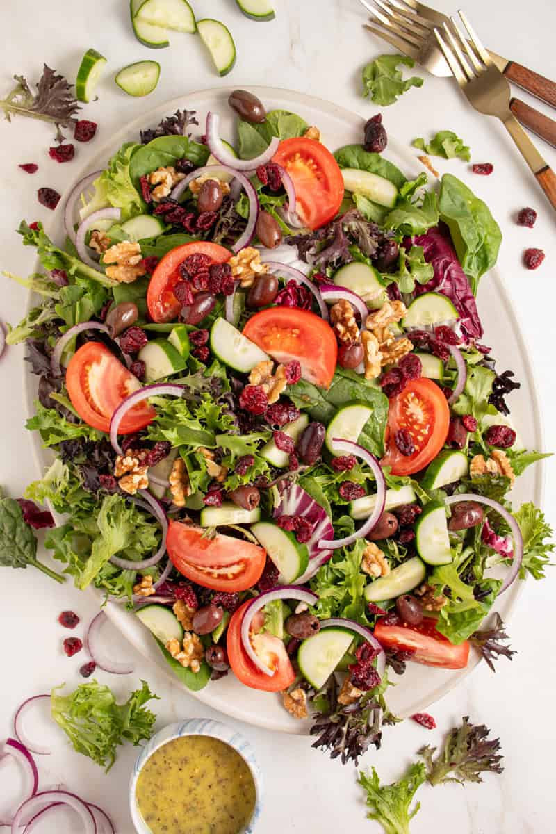 A large platter full of spring mix salad with tomatoes, cucumbers, red onion, walnuts, dried cranberries, and olives.