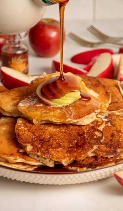 A drizzle of maple syrup being pored onto a stack of apple pancakes.