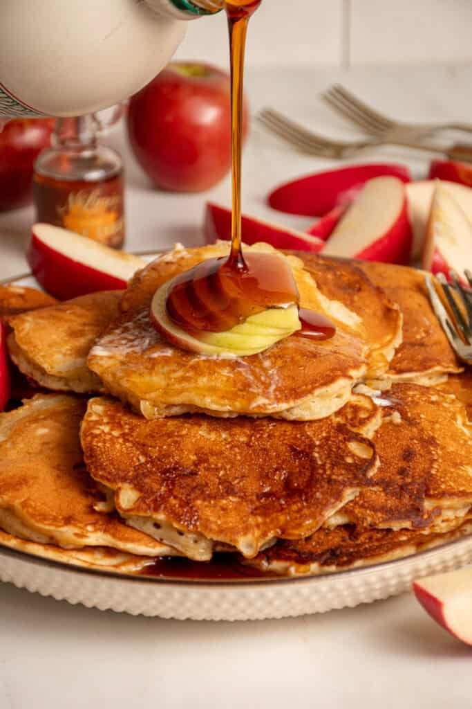 A drizzle of maple syrup being pored onto a stack of apple pancakes.