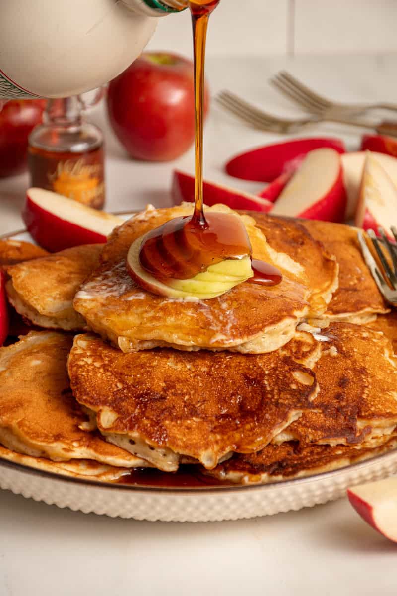 A drizzle of maple syrup being pored onto a stack of apple pancakes.