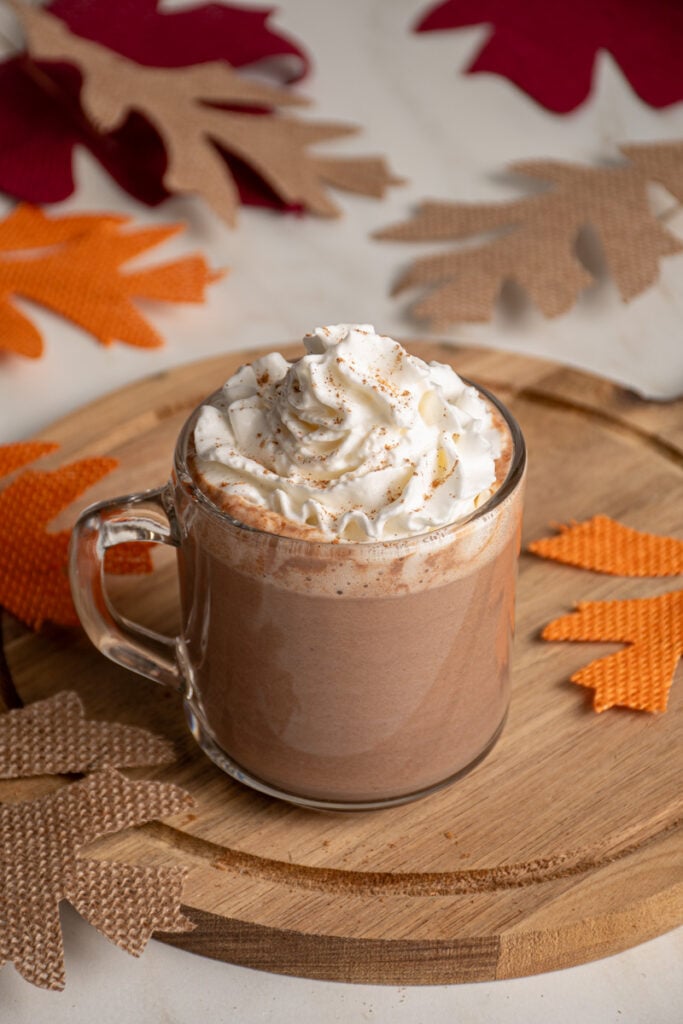 A clear glass mug with pumpkin hot chocolate and topped with whipped cream.
