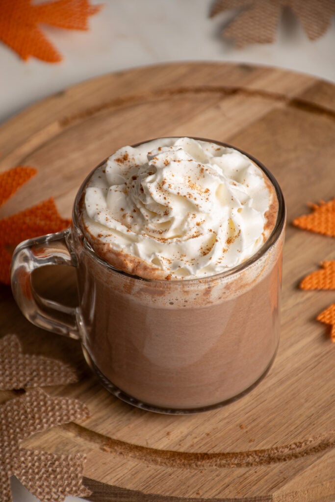 A clear glass mug with pumpkin hot chocolate and topped with whipped cream.