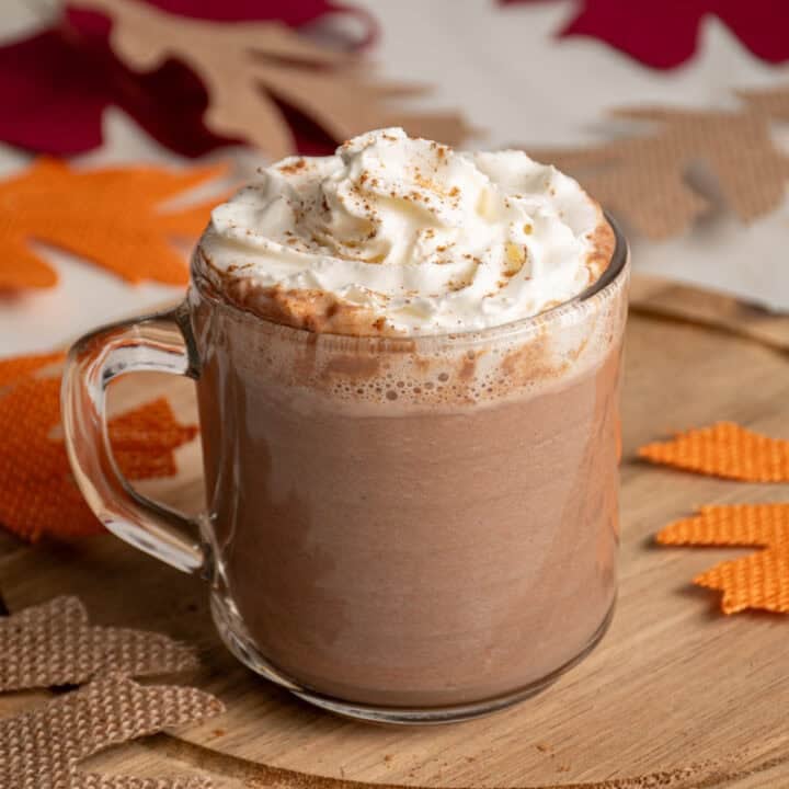 A clear glass mug with pumpkin hot chocolate and topped with whipped cream.
